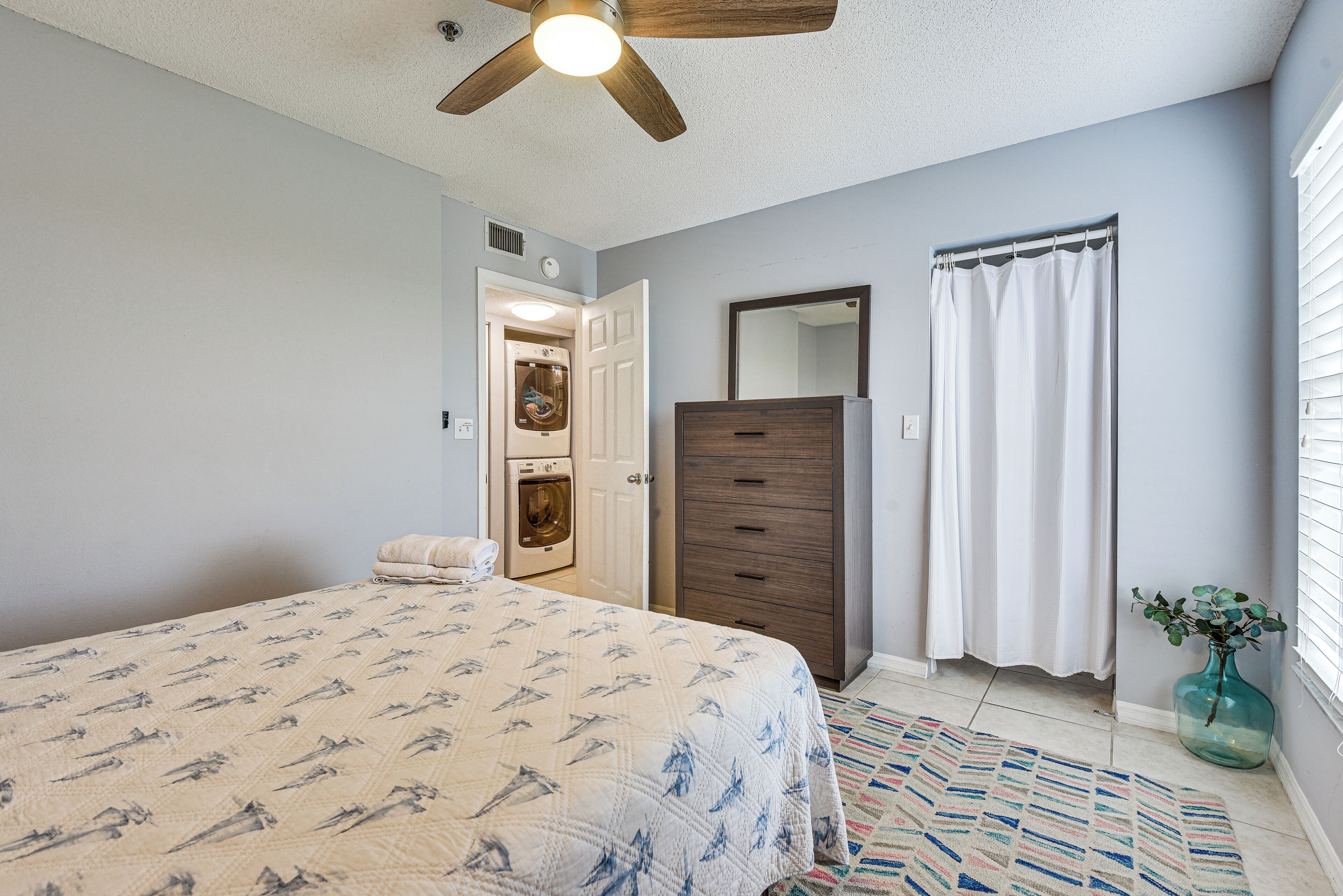 Un dormitorio con cama, cómoda, espejo, ventilador de techo y ventana con cortina.
