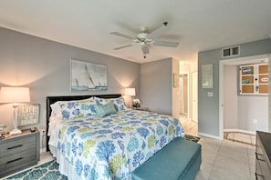Quarto com cama, ventilador no teto, mesa de cabeceira, luminária, um banquinho e porta que dá para outro cômodo.