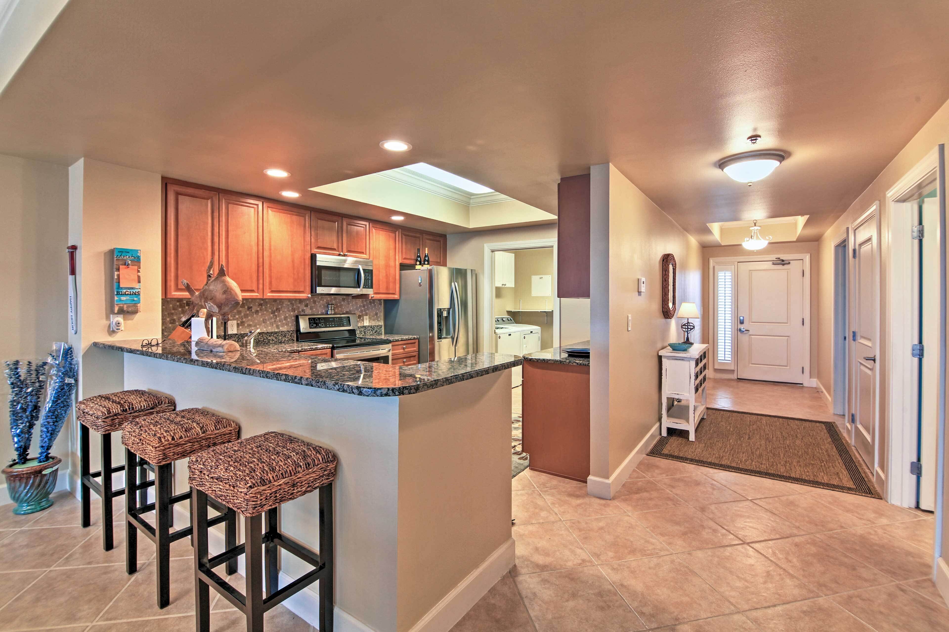 A modern kitchen with wooden cabinets, a granite countertop, and a built-in microwave. There are three wicker bar stools and a small island with a sink.