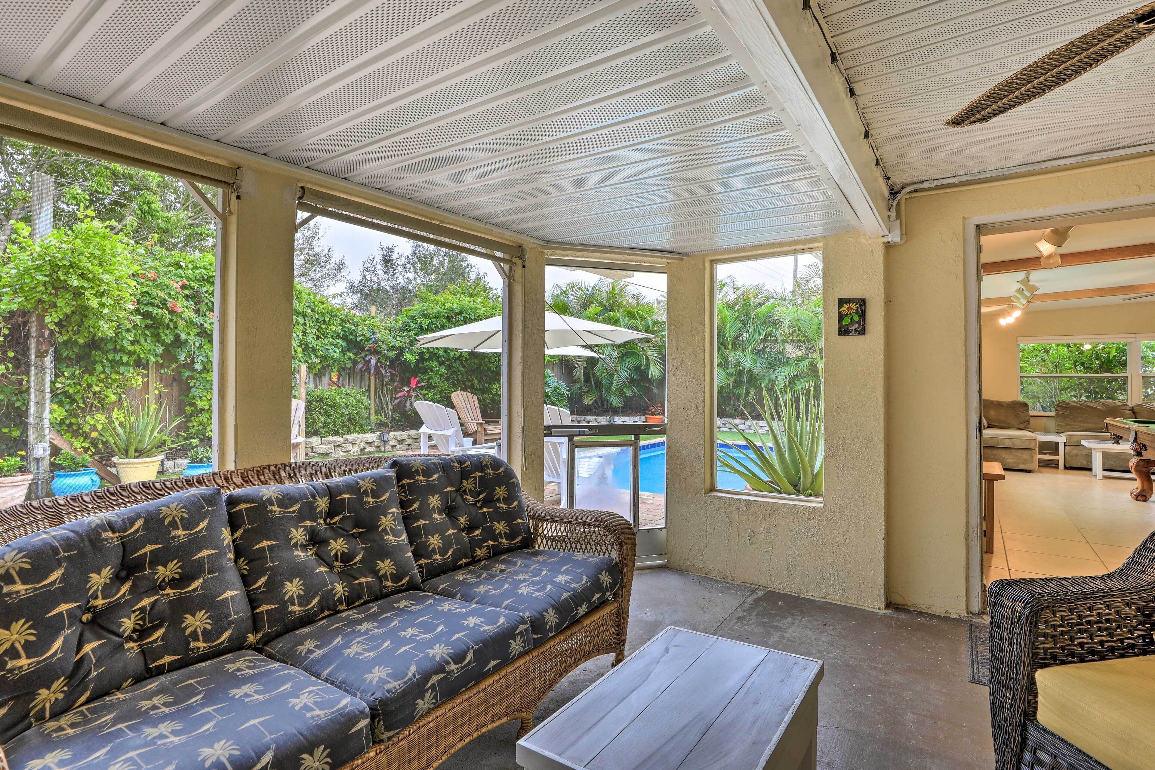 Un espace extérieur aménagé, abrité et doté de mobilier en osier, de grandes fenêtres et d’une vue sur une piscine.
