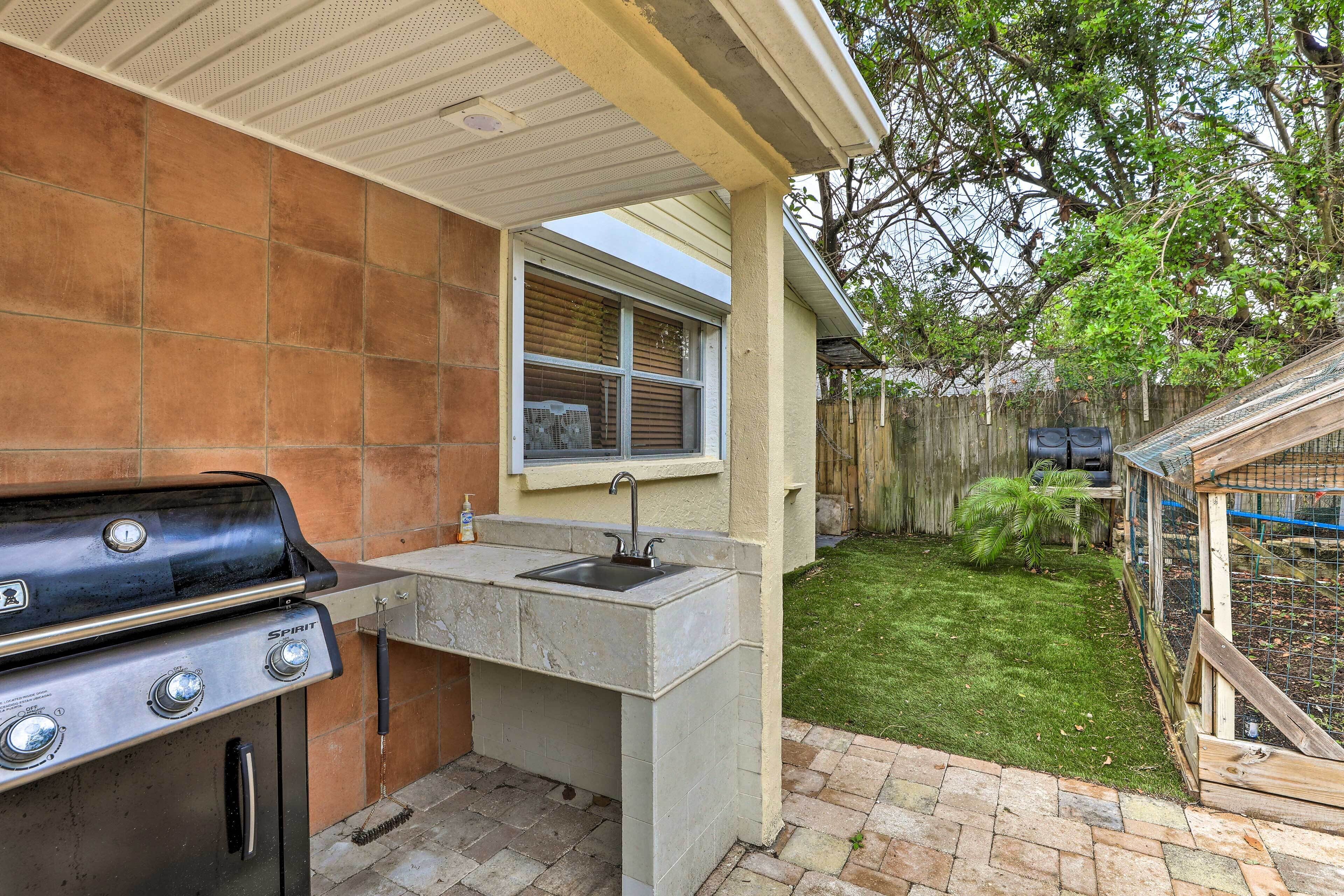 Una cocina exterior con una parrilla, un lavadero y una ventana con vista a un patio trasero con cerca de madera y un árbol.