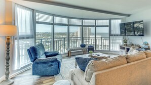 A spacious living room with a large window offering a city view, a comfortable seating area with sofas, a flat-screen TV mounted on the wall, and a small side table with decorative items. - Beautiful Daytona Beach Shores Condo w/ Hot Tub! (Daytona Beach Shores)
