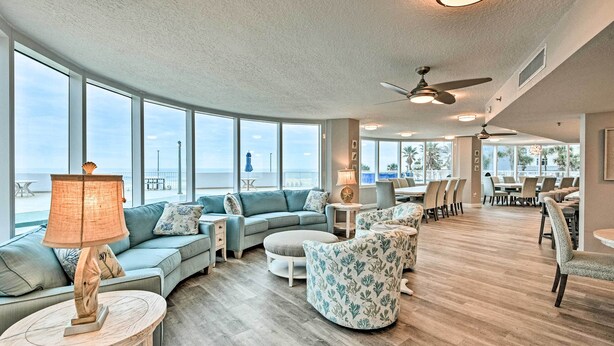 A spacious lounge area with a large window overlooking the beach, a curved sofa, round tables, and chairs arranged for dining. - Beautiful Daytona Beach Shores Condo w/ Hot Tub! (Daytona Beach Shores)