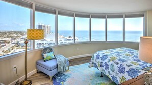 A bedroom with a large bay window, a bed with a floral bedspread, a floor lamp, and a view of the ocean and cityscape. - Beautiful Daytona Beach Shores Condo w/ Hot Tub! (Daytona Beach Shores)