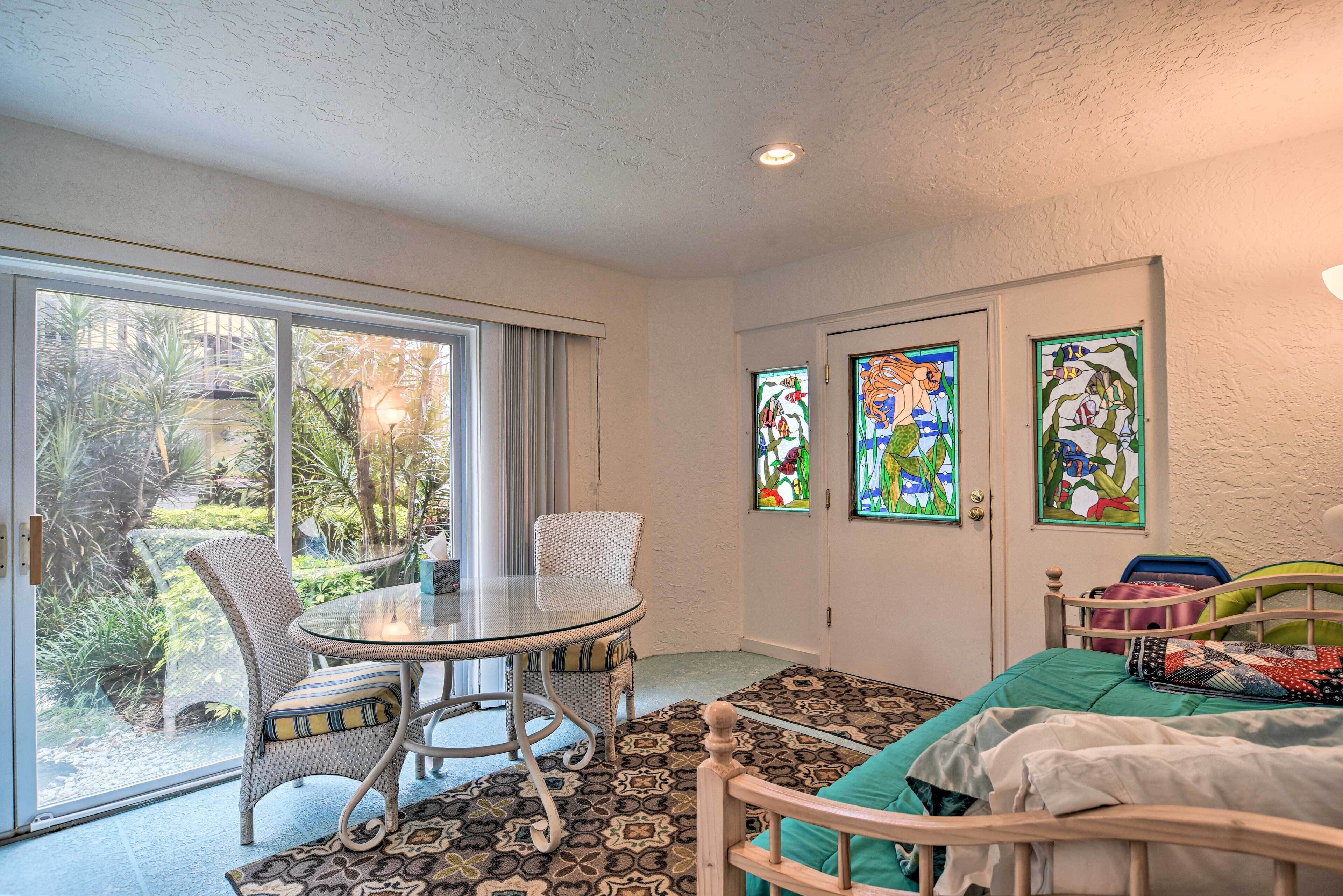 A room with a glass door leading to a garden, a round glass table with wicker chairs, a bed with a patterned bedspread, and two stained glass windows.