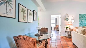 Un comedor con mesa de vidrio y sillas de mimbre, una sala de estar con sofá y lámpara, y un pasillo con puerta que da a otra habitación.