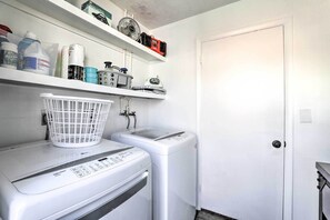 A laundry room with a front-loading washer and dryer, a white basket on the washer, and shelves above holding various items. - Beachy Bradenton Vacation Home Near Ocean! (Bradenton)