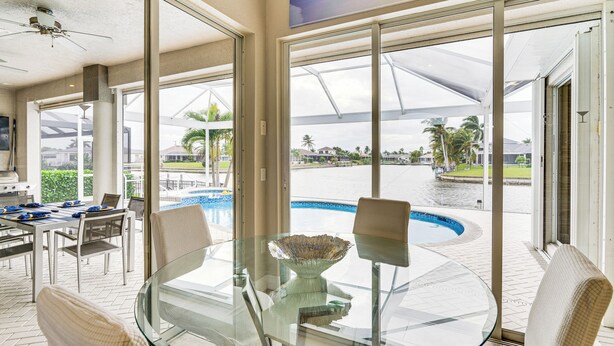 Mesa de jantar com tampo de vidro, cadeiras, uma tigela e vista para uma piscina e palmeiras.