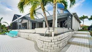 Uma casa moderna com fachada de vidro, um muro de pedra de contenção e área de piscina com cadeiras azuis.
