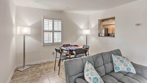 A living room with a gray sofa, two floor lamps, a dining table with chairs, and a window with blinds.