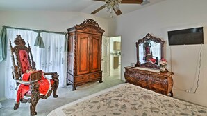 A bedroom with a large bed, a wooden wardrobe, a red upholstered chair, a ceiling fan, and a television mounted on the wall. - Bright Spring Hill Home: 5 Mi to Weeki Wachee Park (Spring Hill)