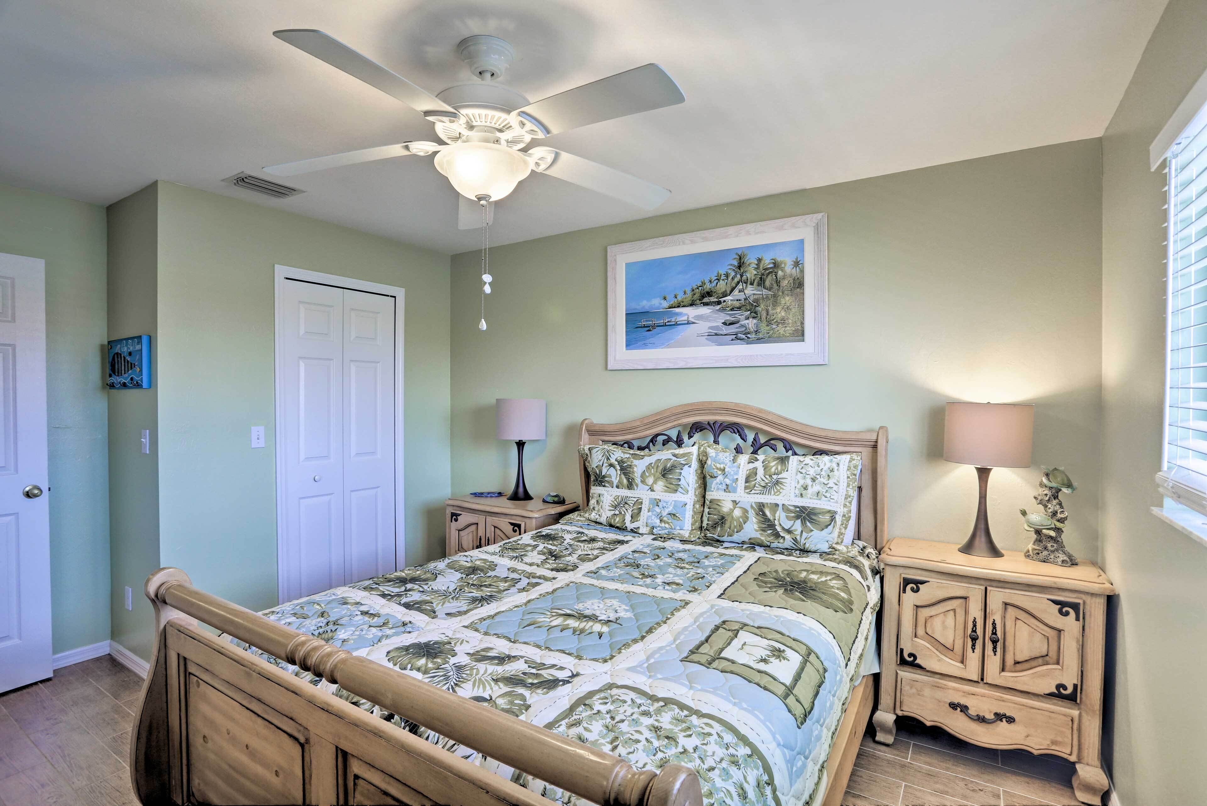 A bedroom with a bed, a ceiling fan, a nightstand, a lamp, a door, and a framed picture on the wall.