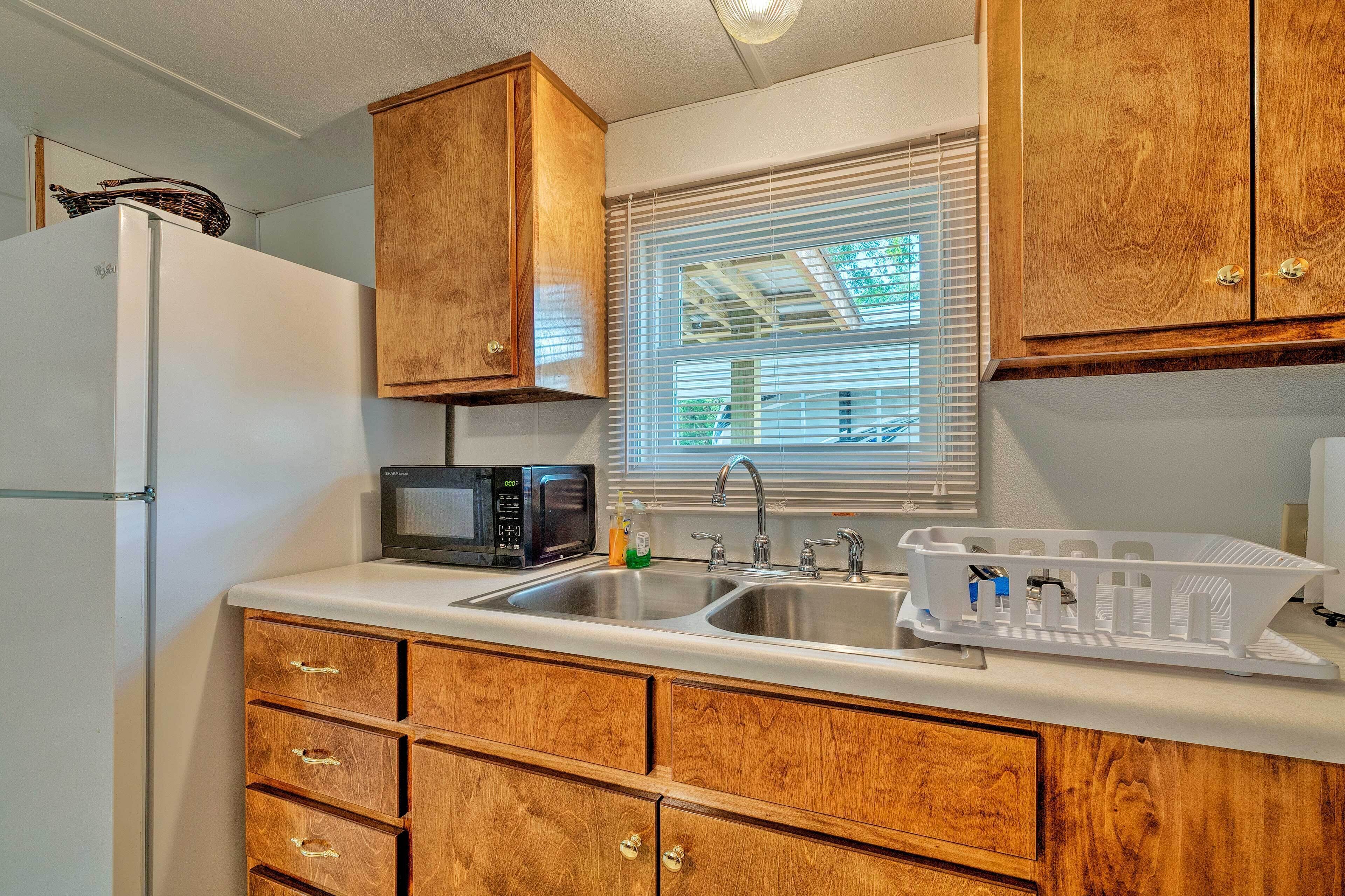 A kitchen with wooden cabinets, a white refrigerator, a microwave, a sink with a dish rack, and a window with blinds.