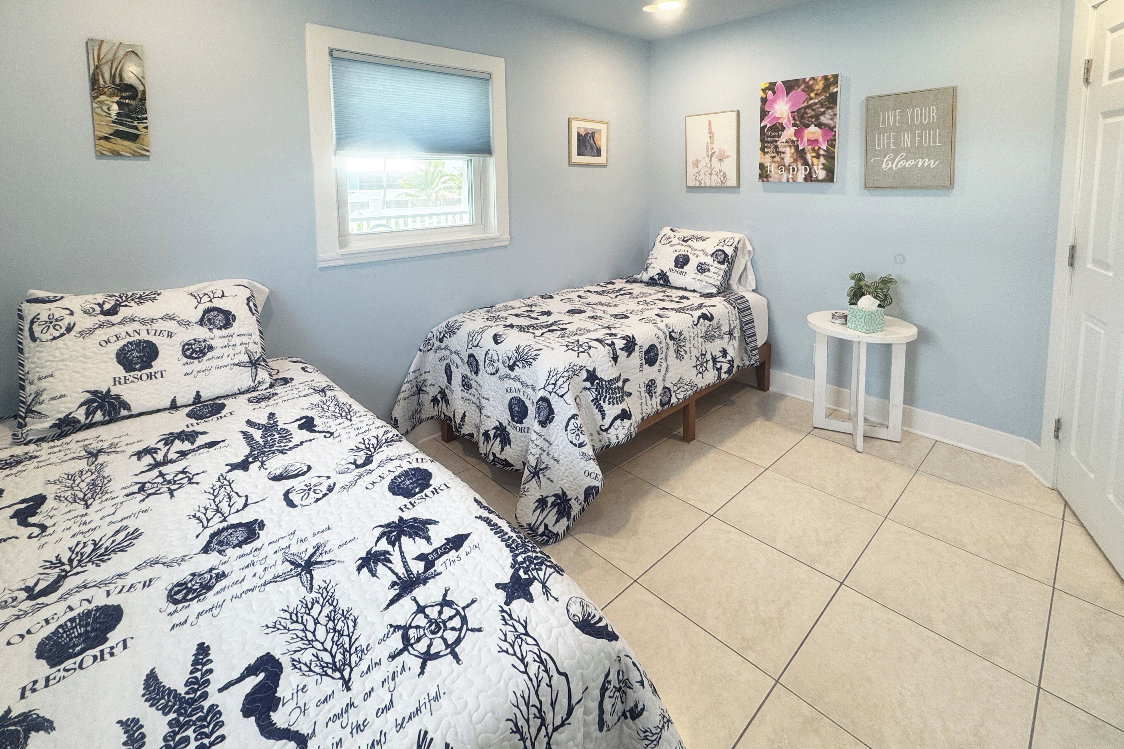 A bedroom with two beds, a window, a small table with a plant, and wall art.
