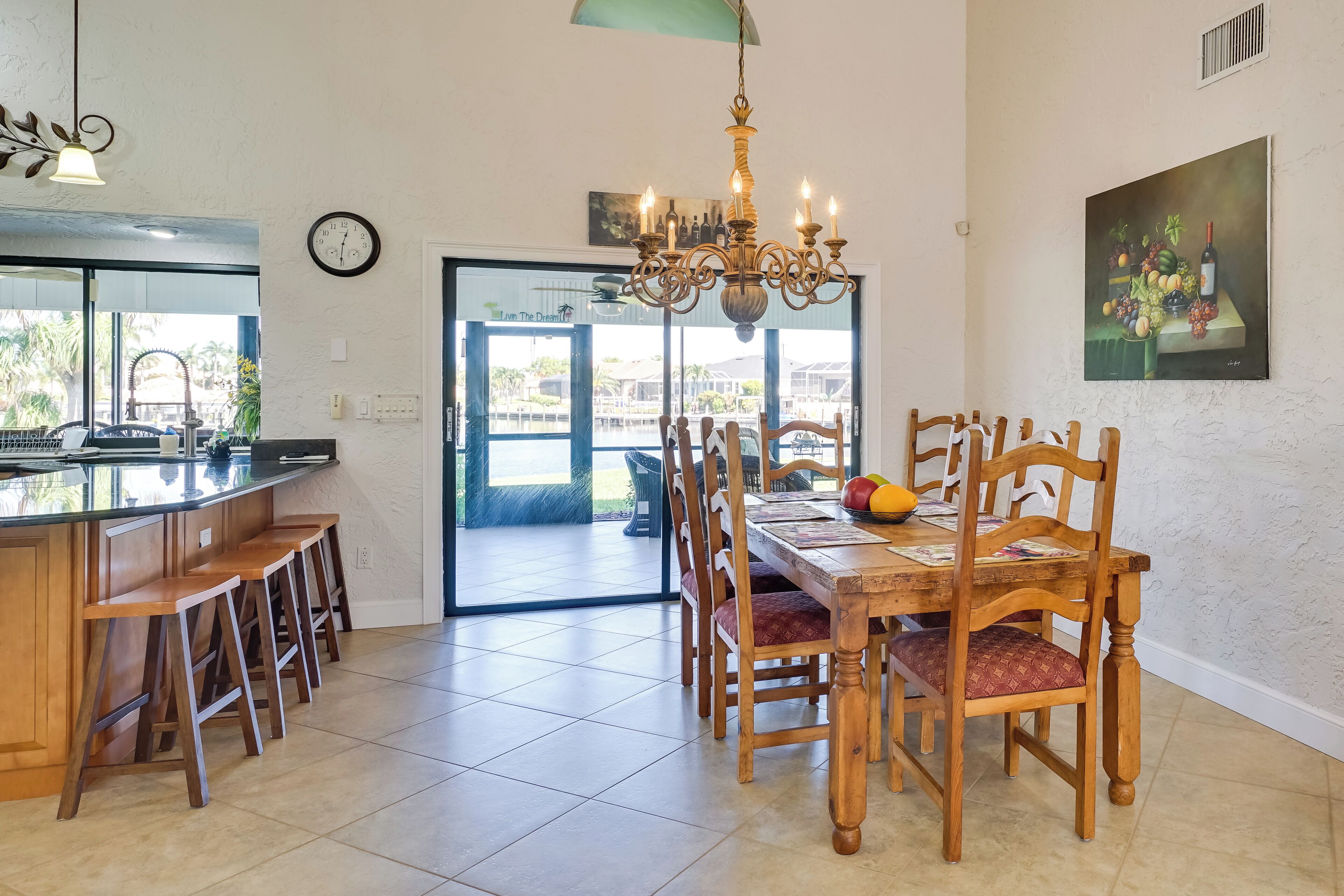 Une cuisine spacieuse avec un coin bar, des chaises en bois, un lustre et une table à manger agrémentée d’un panier de fruits.