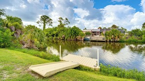 A lakeside property with a dock, lush greenery, and a house in the background. - Canal-front Retreat w/ Heated Pool & Hot Tub! (Port Charlotte)