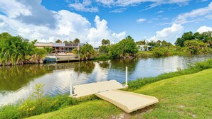 A riverside view with a dock, grassy banks, and buildings in the background. - Canal-front Retreat w/ Heated Pool & Hot Tub! (Port Charlotte)
