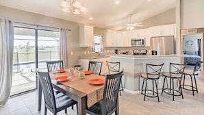 A modern kitchen with white cabinets, a central island with bar stools, and a dining area with a table set for four. - Central Cape Coral House w/ Private Screened Pool! (Cape Coral)