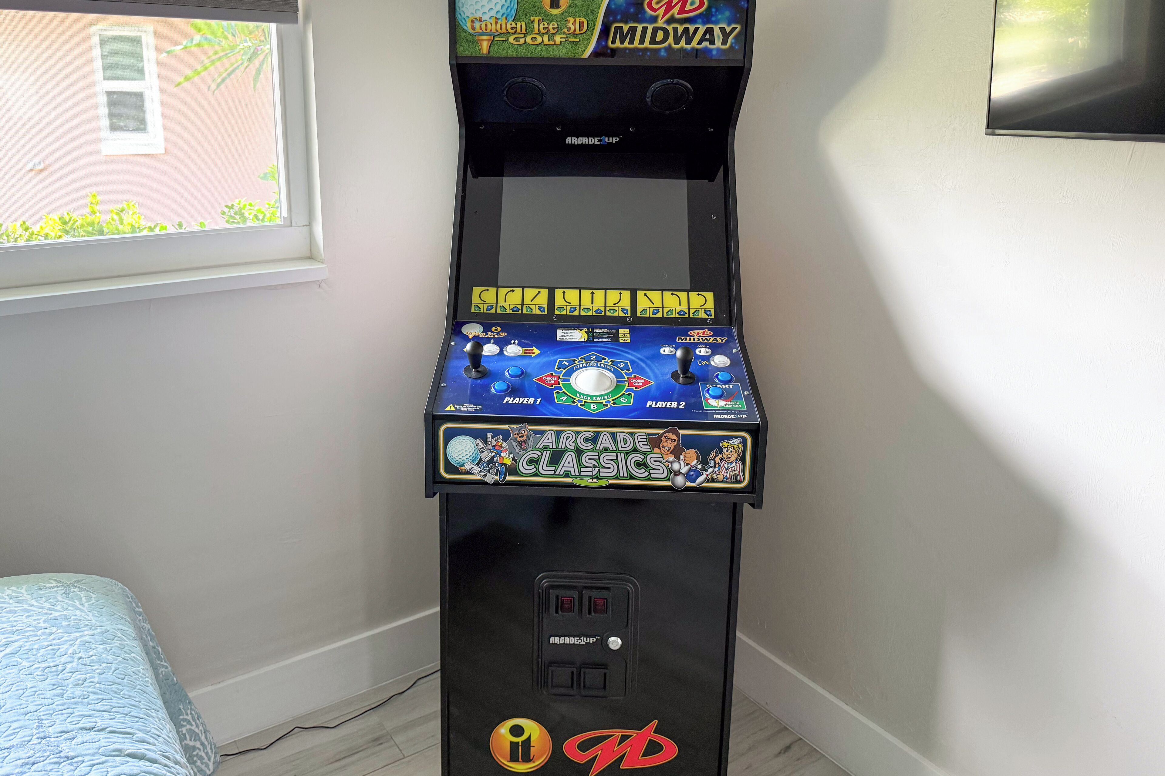 Une borne d’arcade noire, dotée d ’un écran et de boutons, est placée dans un coin d’une pièce qui contient une fenêtre et un lit.