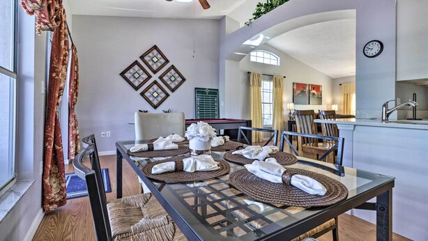 A dining area with a glass table set for four, surrounded by chairs. A ceiling fan is visible above, and there are decorative wall art pieces. - Charming Kissimmee Retreat w/ Pool & Lanai! (Kissimmee)
