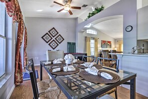 Uma área de refeições com mesa de vidro posta para quatro pessoas, cercada por cadeiras. Um ventilador de teto é visível acima, e há peças de arte decorativas nas paredes.