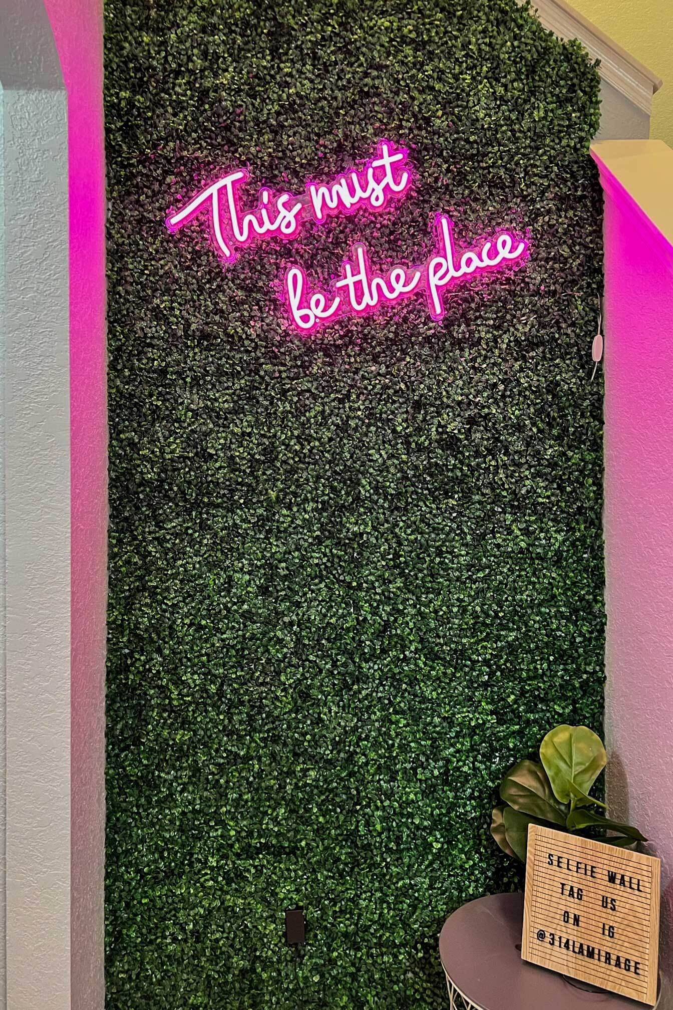 A neon sign with the text "This must be the place" against a backdrop of green foliage.