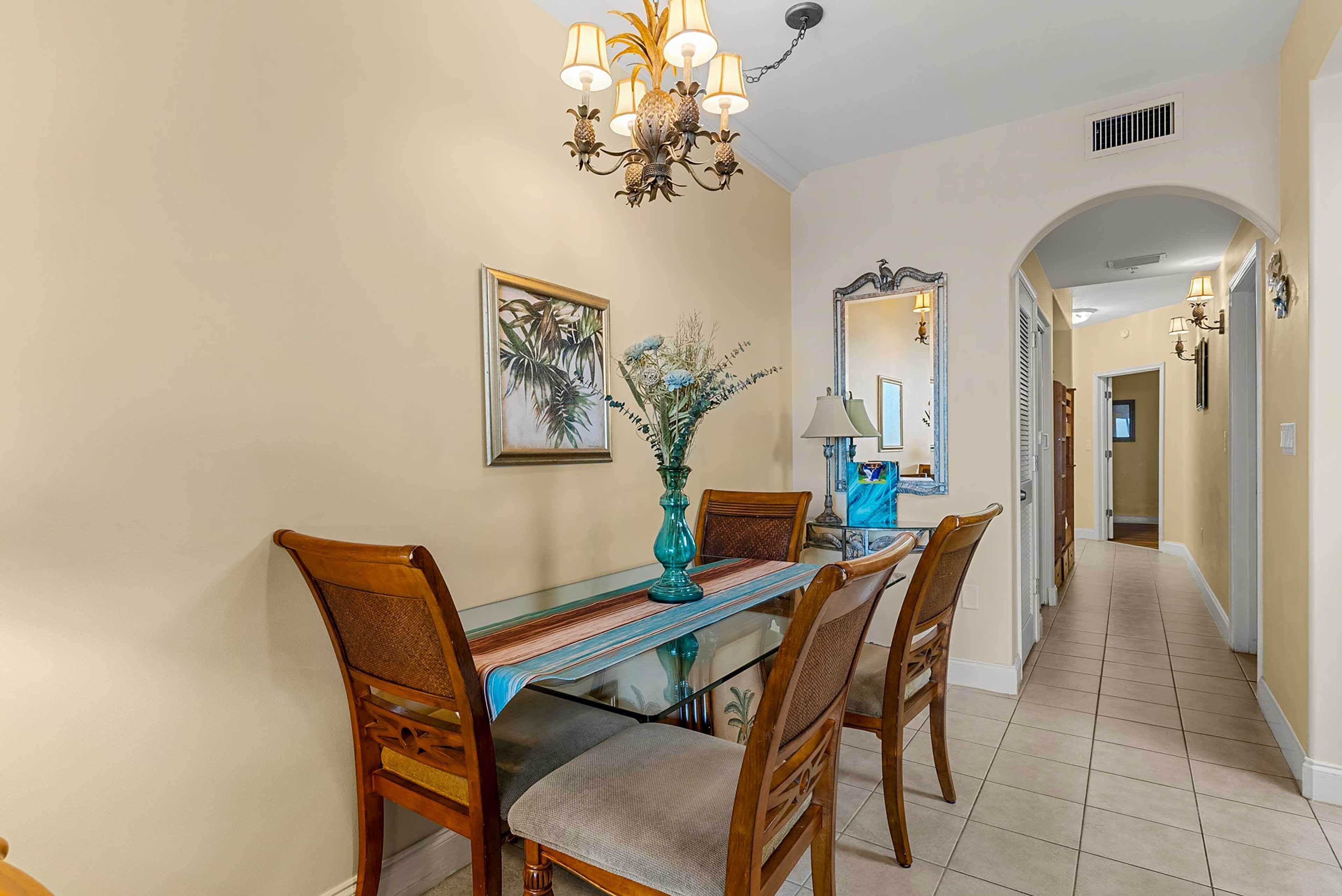 A dining area with a glass table, wooden chairs, a vase with flowers, a chandelier, and a mirror on the wall.