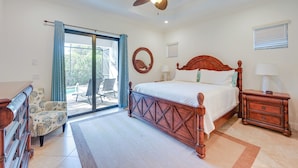 Ein Schlafzimmer mit einem großen Bett, einem Deckenventilator, einem Nachttisch, einem Sessel und Blick durch eine Glastür.