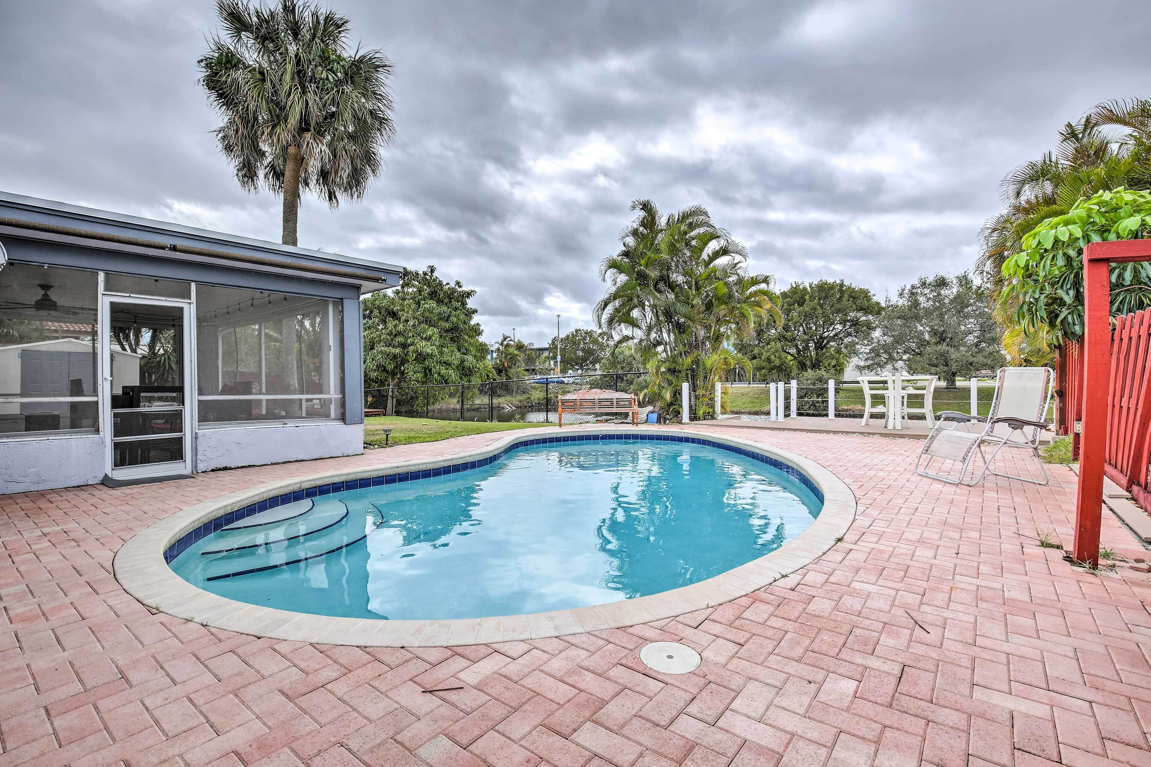 A swimming pool with a brick patio, a covered area with glass doors, and palm trees in the background.