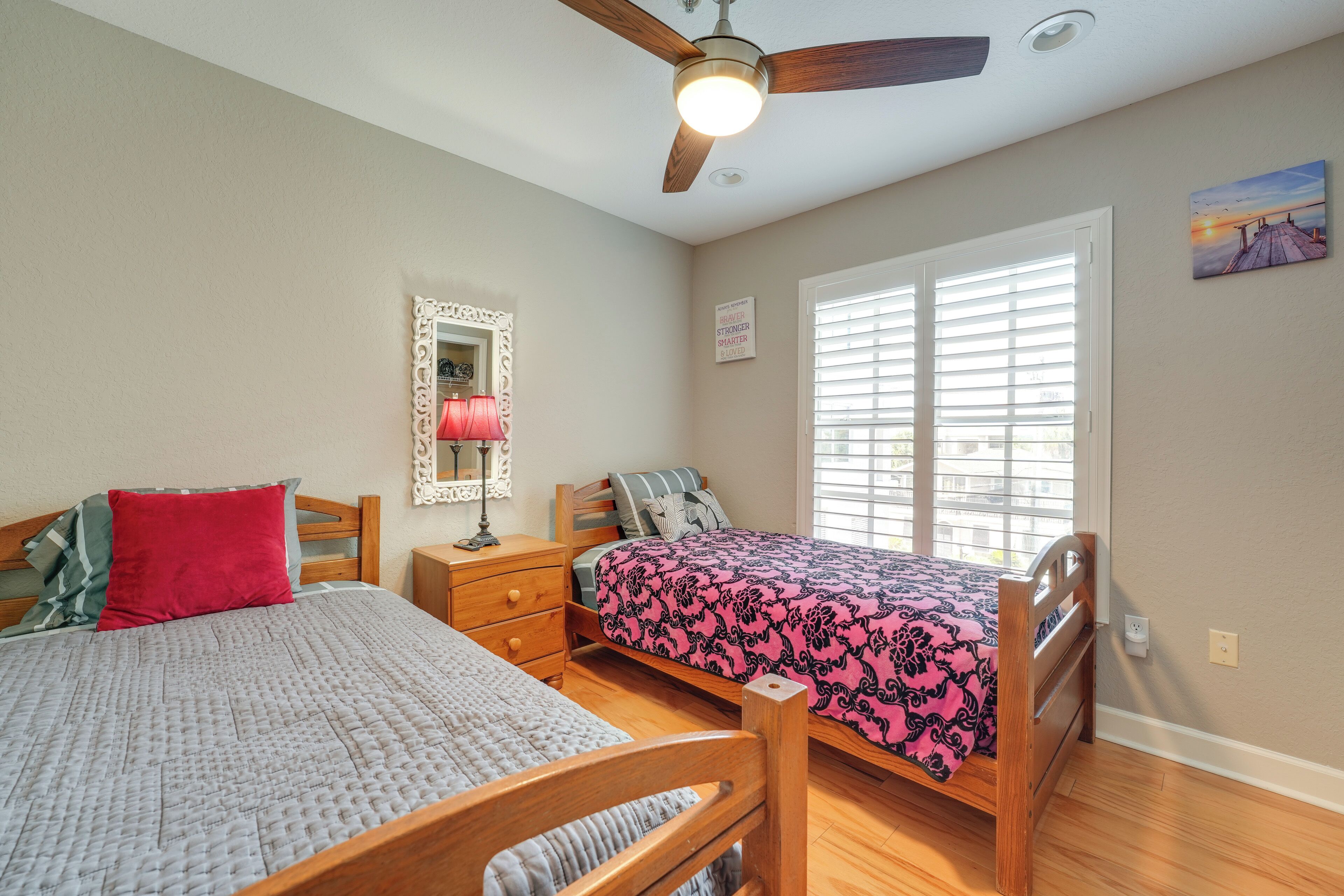 A bedroom with two beds, a ceiling fan, a window with blinds, a nightstand, and a wall-mounted mirror.
