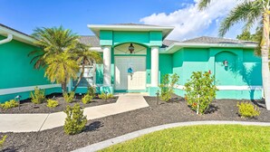 A turquoise house with a white door, surrounded by a garden with small plants and a palm tree. - Colorful Cape Coral Vacation Rental w/ Pool! (Cape Coral)