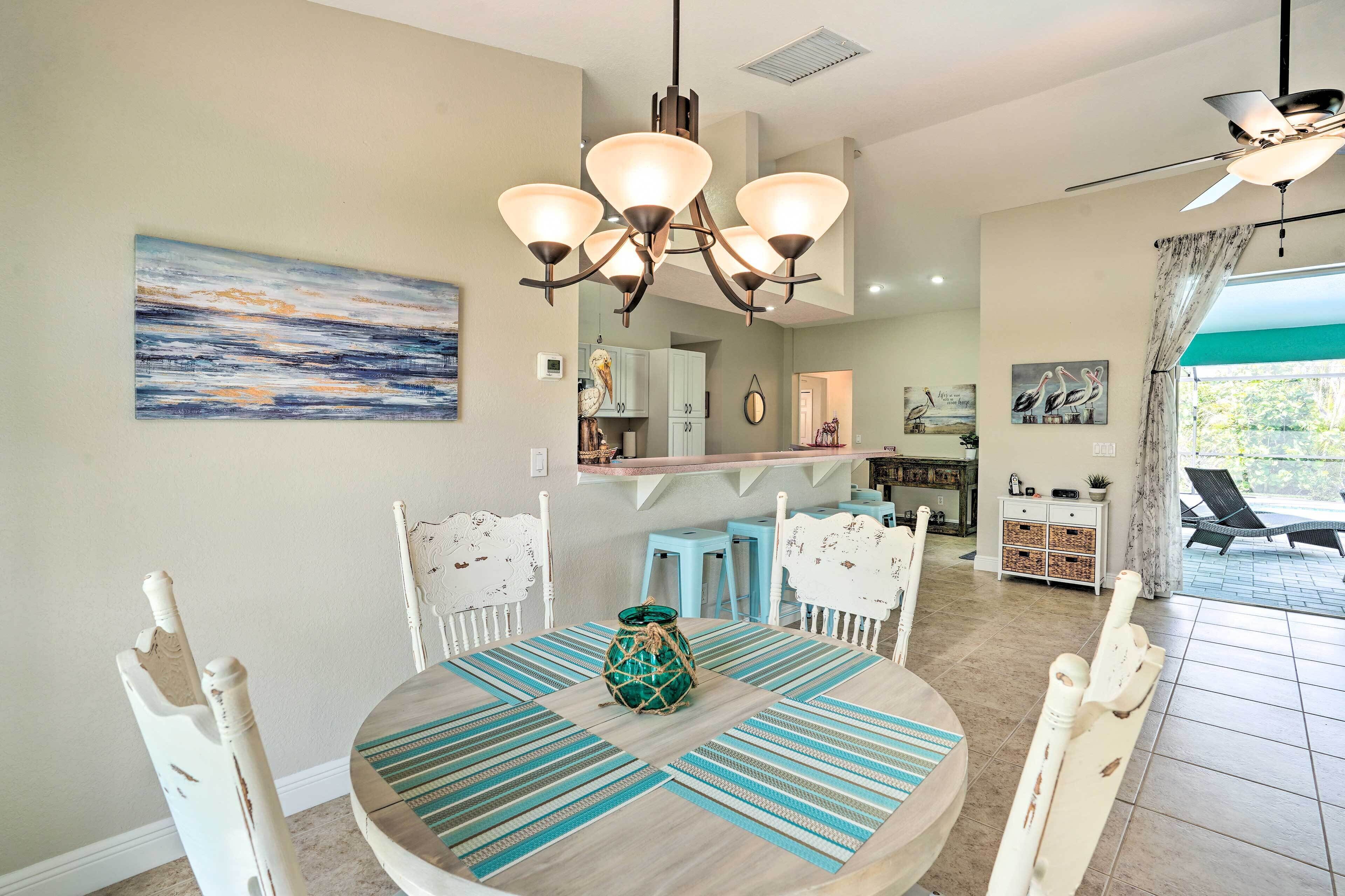 A dining area with a round table, white chairs, and a chandelier. There is a kitchen in the background with a counter and stools.