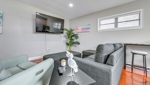 Un salon moderne avec un canapé gris, un fauteuil bleu clair, une petite table avec une télécommande, un oiseau décoratif et un téléviseur fixé au mur.