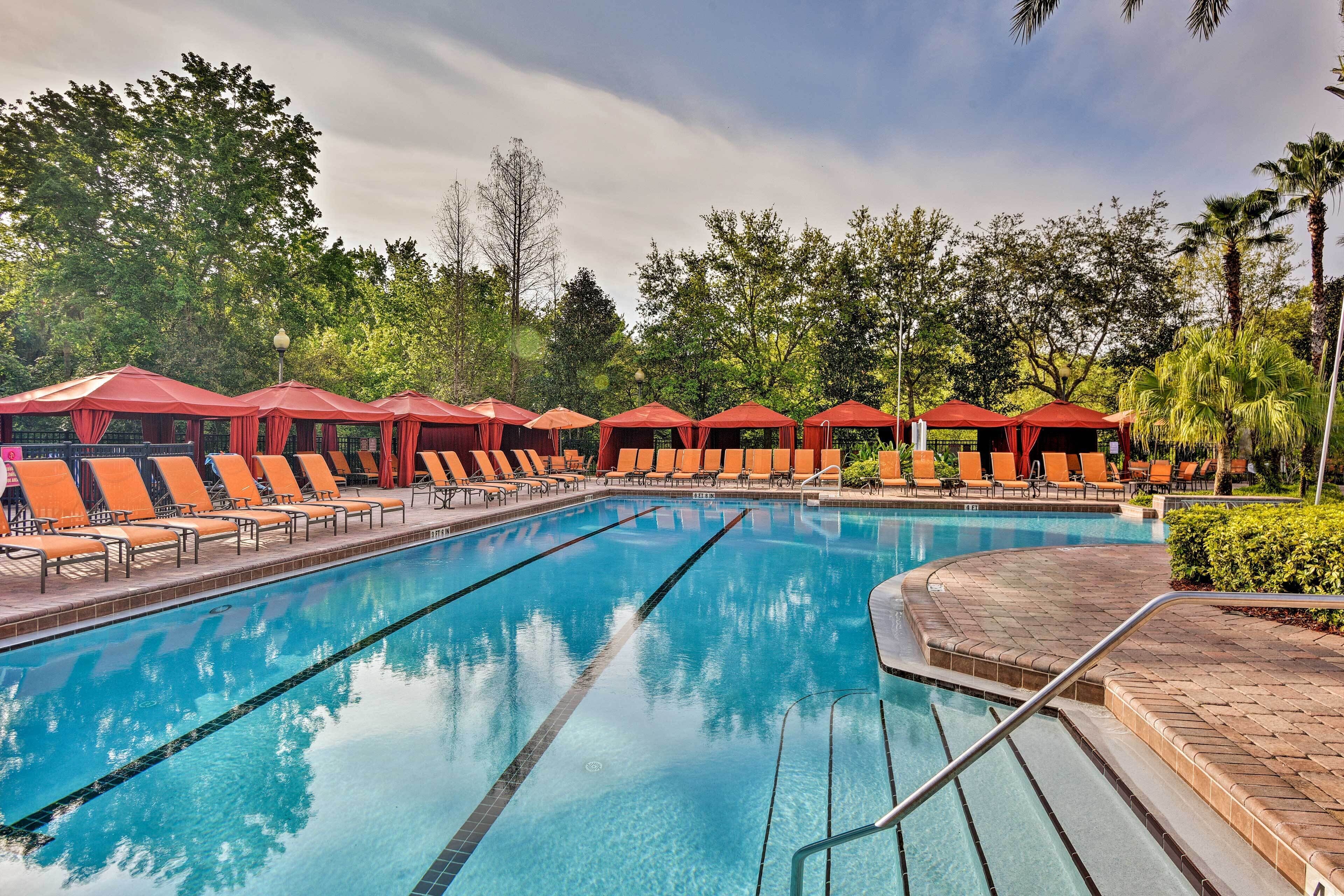 A swimming pool with multiple lanes, surrounded by orange lounge chairs and red umbrellas, set in a lush, green environment.