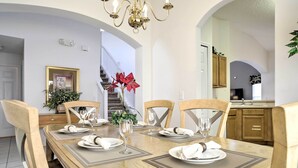 Une salle à manger avec une table en bois dressée pour quatre personnes, des chaises, un lustre et un escalier en arrière-plan.