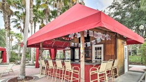 Un espace bar extérieur doté de parasols rouges, d’un comptoir en bois et de nombreux tabourets de bar.