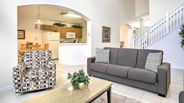 Un salon comprenant un canapé, un fauteuil et une table basse. On y trouve un escalier avec une rampe et une porte qui donne sur une autre pièce.