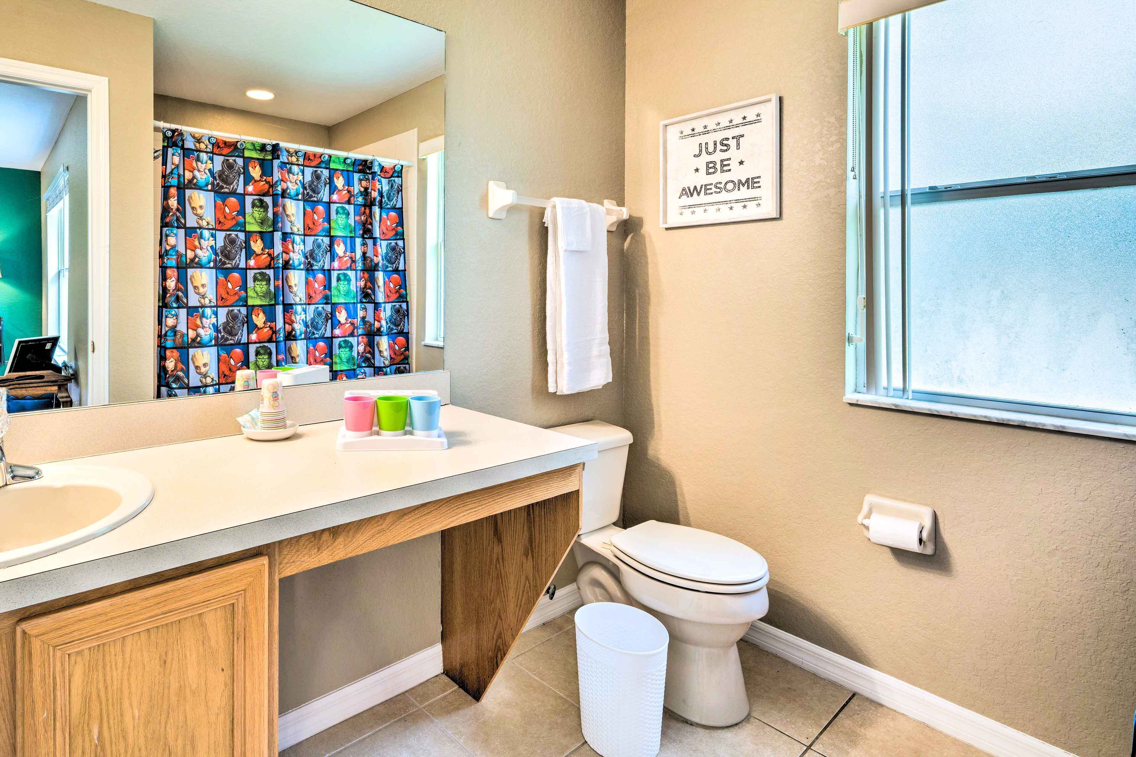 A bathroom with a toilet, sink, and a shower curtain with superhero characters. There is a sign on the wall and a window.
