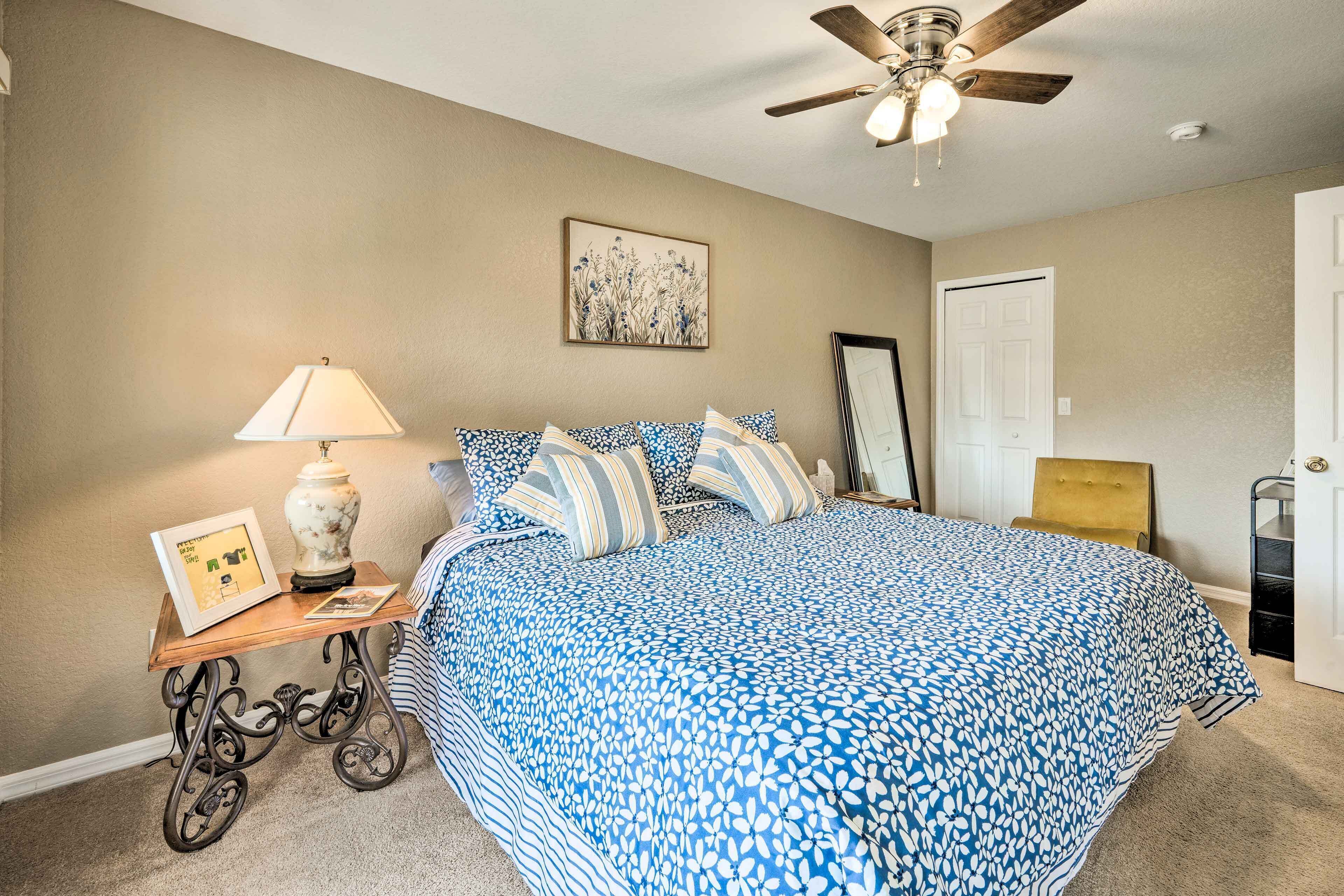 A bedroom with a large bed, a ceiling fan, a bedside table with a lamp, and a framed picture.