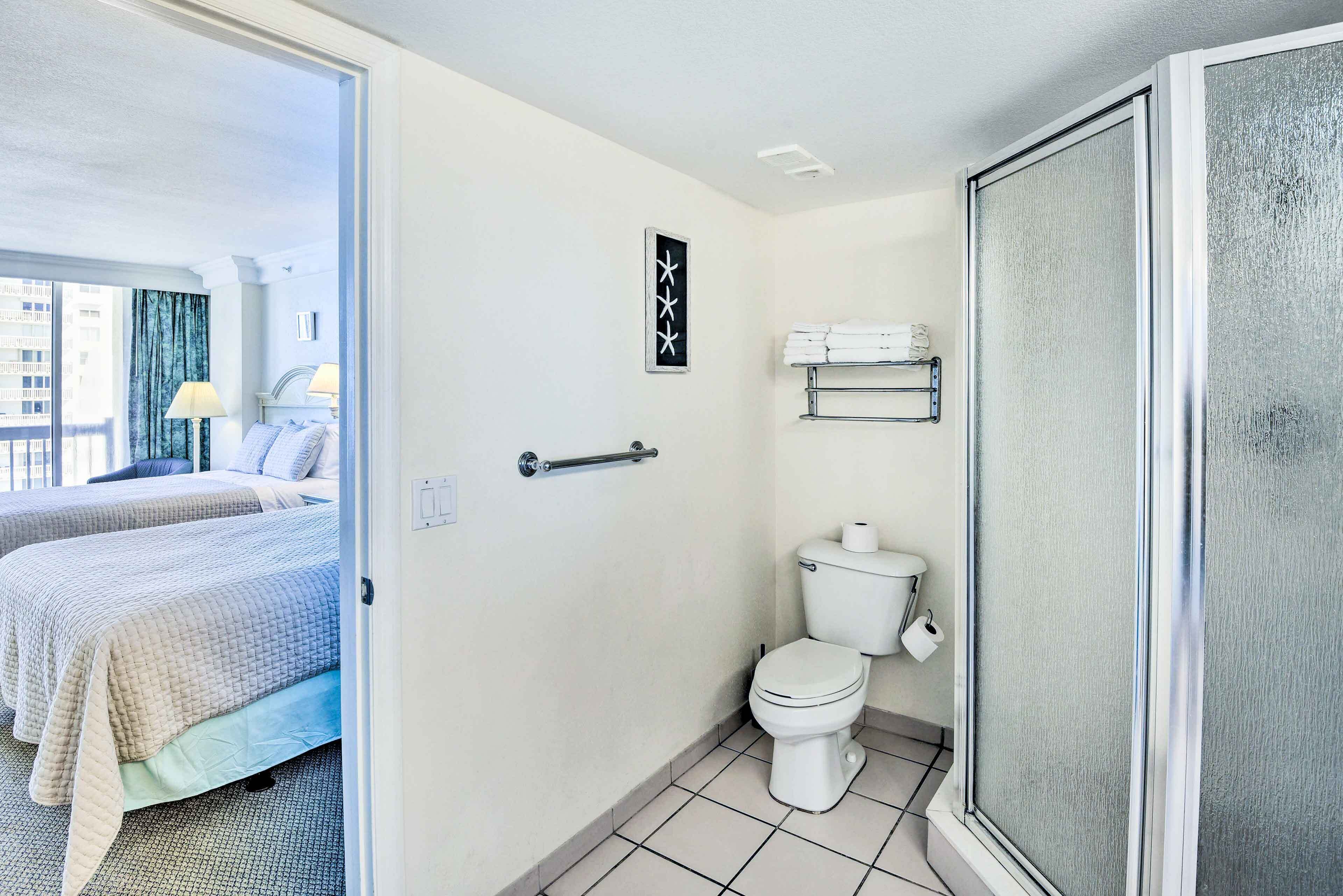 Une salle de bain d’hôtel avec des toilettes, une douche et un lit visible à travers une porte ouverte.