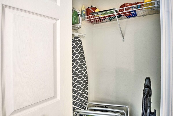 Casa de banho pequena e estreita com porta branca, um porta-toalhas com uma toalha estampada, uma prateleira com produtos de limpeza e um aspirador.