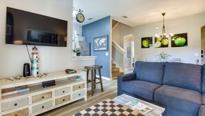 Una sala de estar con un televisor de pantalla plana grande, un sofá azul, una mesa de centro con mantel individual a cuadros y una estantería con libros y objetos decorativos.