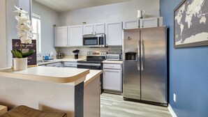 Una cocina moderna con armarios blancos, un refrigerador de acero inoxidable, un microondas y un tostador. Hay una pequeña isla con una planta y un letrero que dice 'sean nuestros huéspedes'.
