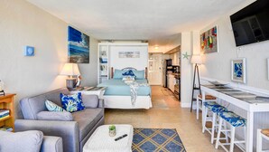 A compact living space with a sofa, a bed, a small table, a bar area, and a kitchen in the background. - Daytona Beach Studio With Balcony + Ocean View (Daytona Beach Shores)
