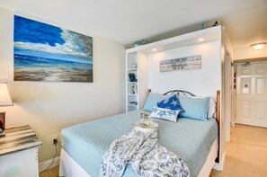 Une chambre à coucher avec un lit, un tableau représentant l’océan, une étagère avec des objets décoratifs et un panneau accroché au mur.