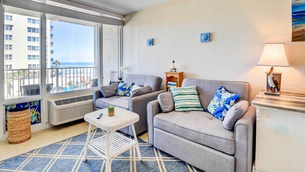 A living room with two grey sofas, a small white table, and a view of the ocean through large windows. - Daytona Beach Studio With Balcony + Ocean View (Daytona Beach Shores)