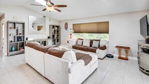 Ein geräumiges Wohnzimmer mit Deckenventilator, großem Fenster mit Jalousien, Fernseher und Bücherregal.