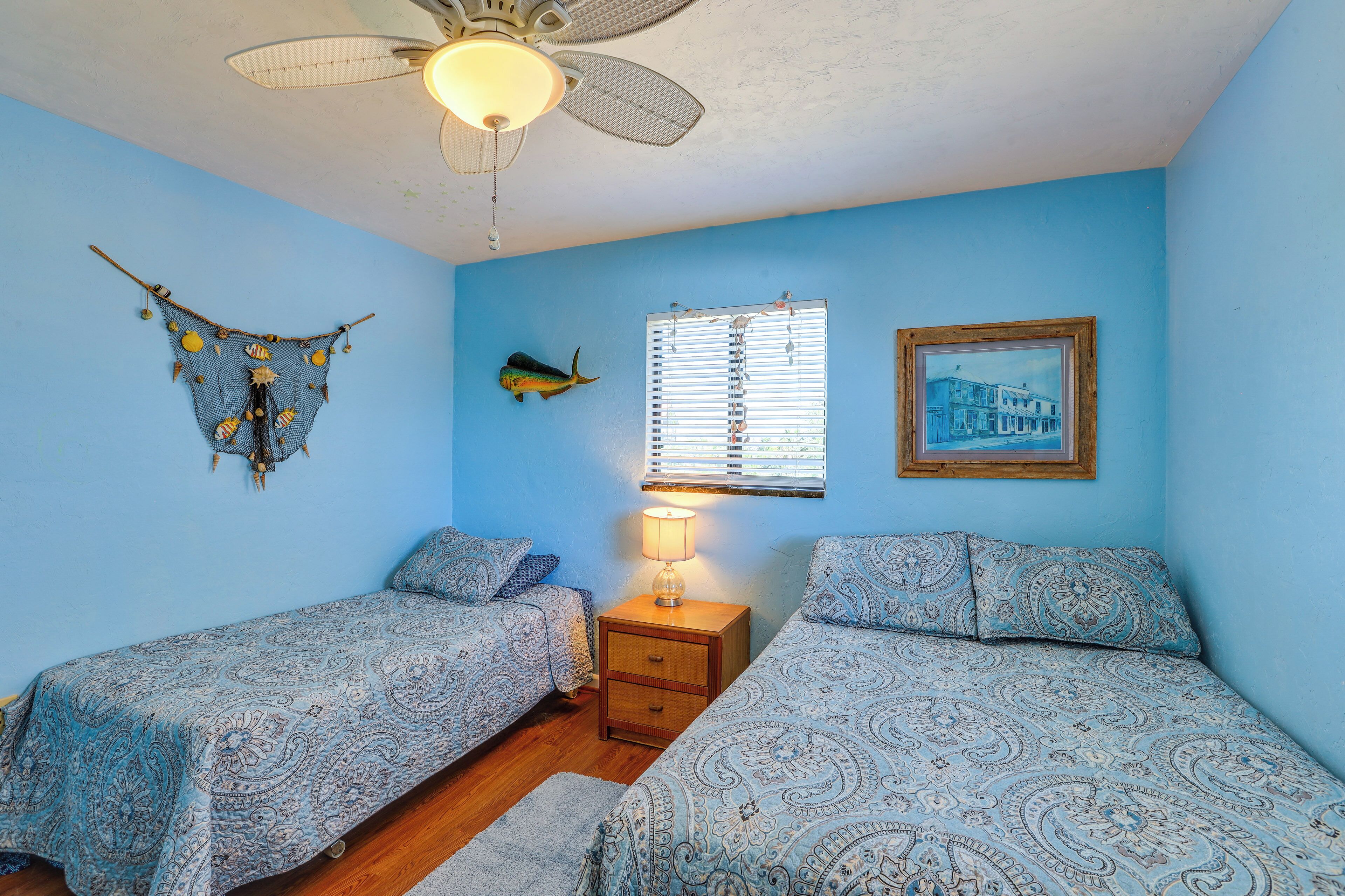 A room with two beds, a ceiling fan, a wall-mounted fish decoration, a window with blinds, and a framed picture.