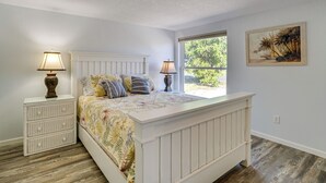 A bedroom with a large bed, two bedside tables with lamps, a window with a view of greenery, and a painting on the wall.
