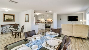 A dining area with a glass table, chairs, and a blue placemat. There is a kitchen in the background with white cabinets and a TV mounted on the wall.